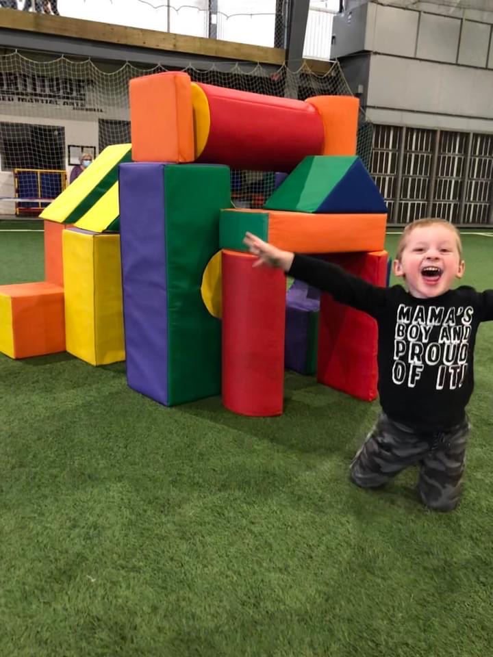child smiling with arms wide in front of structure of blocks behind them.